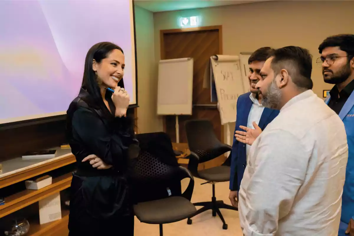 Un grupo de personas conversando en una sala de conferencias, con una mujer sonriendo mientras sostiene un bolígrafo y tres hombres escuchándola atentamente.